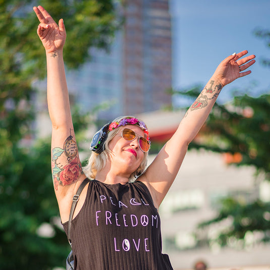 Quiet Events Silent Disco headphones for property management. A young woman outdoors enjoying her time in the sun with her hands up in the air wearing headphones.